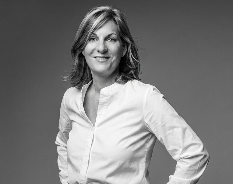 Frau mit lockigem Haar in einem weißen Hemd und schwarzer Hose, lächelt selbstbewusst.